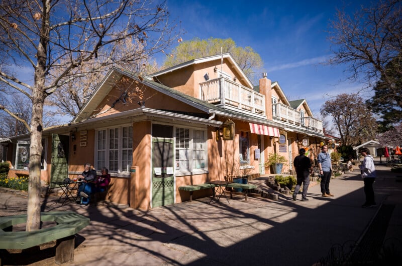 Escena de una calle soleada con gente caminando o sentada afuera de un edificio de estilo adobe color canela con puerta verde, balcón y toldo a rayas. Los árboles desnudos proyectan sombras sobre la acera.