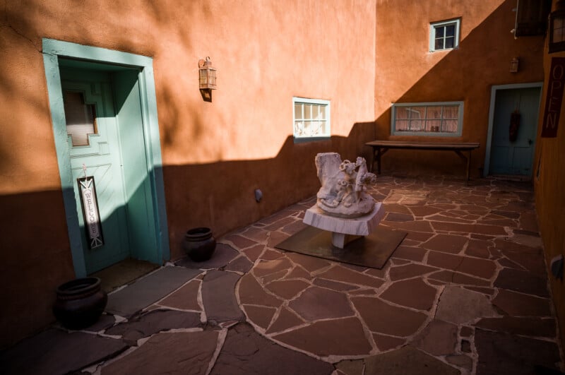 Soleado patio de adobe con paredes de color rojo anaranjado, puerta turquesa, pequeñas ventanas, plantas en macetas y una escultura blanca sobre un pedestal central. Las sombras crean patrones geométricos en el suelo de piedra.