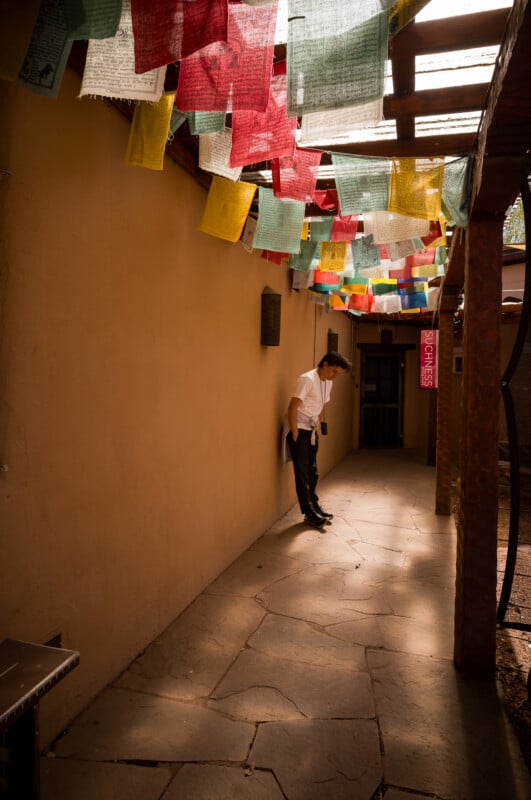 En un estrecho pasillo al aire libre, un hombre vestido con una camisa blanca y pantalones oscuros se apoyaba contra una pared iluminada por el sol de la que colgaban coloridas banderas de oración. La luz se filtra a través del techo de láminas de arriba.