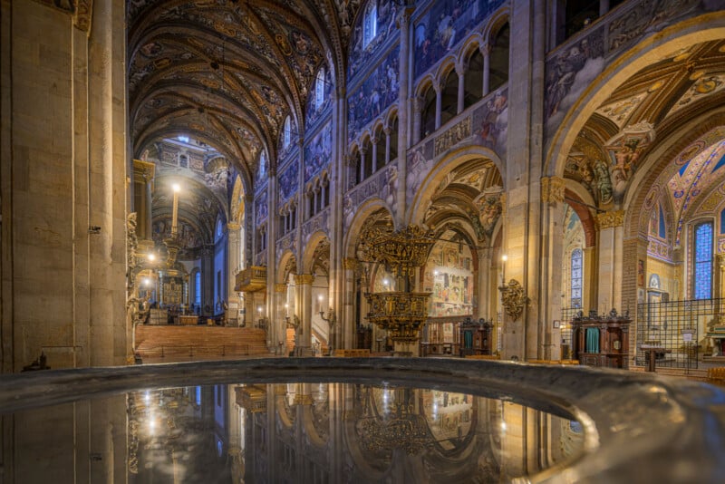 Interior of a grand cathedral with ornate arches, detailed frescoes on the walls and ceiling, and a reflective baptismal font in the foreground, creating a sense of depth and reverence.