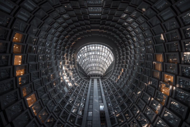 A wide-angle view looking up at a tall, cylindrical building interior with concentric rings of windows, a glass ceiling, and warm light glowing from some windows. The architecture creates a dramatic, tunnel-like effect.