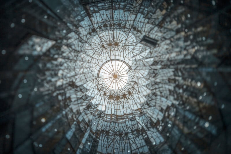 A geometric glass and metal dome ceiling viewed from below, with intricate symmetrical patterns radiating from a central circular skylight, creating a kaleidoscopic effect. The image has a cool, blurred vignette.