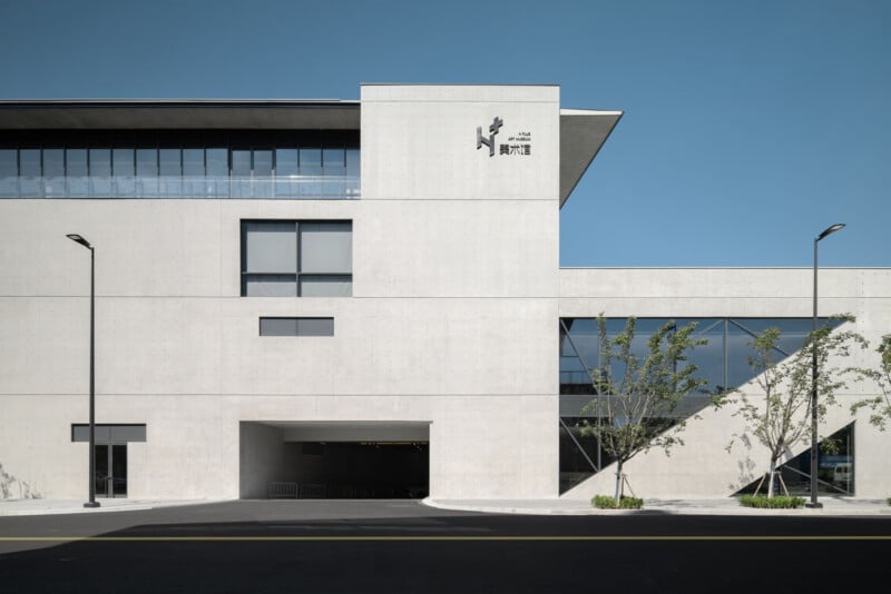 A modern, minimalist white building with large windows, a rectangular entrance, and a few young trees in front. The facade features signage with Chinese characters and a logo near the top. The sky is clear and blue.