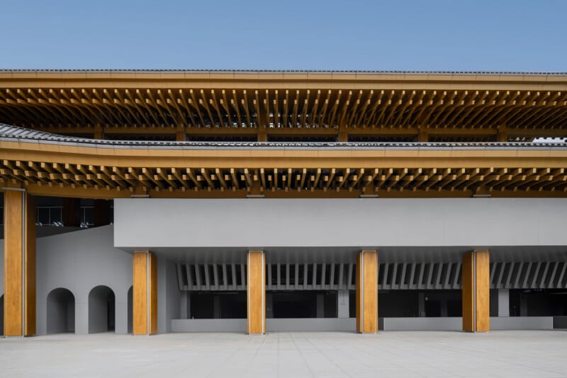 Modern building with a flat roof and multiple layers of exposed wooden beams above a gray concrete facade, featuring tall vertical columns and several open archways at the base. Clear blue sky in the background.