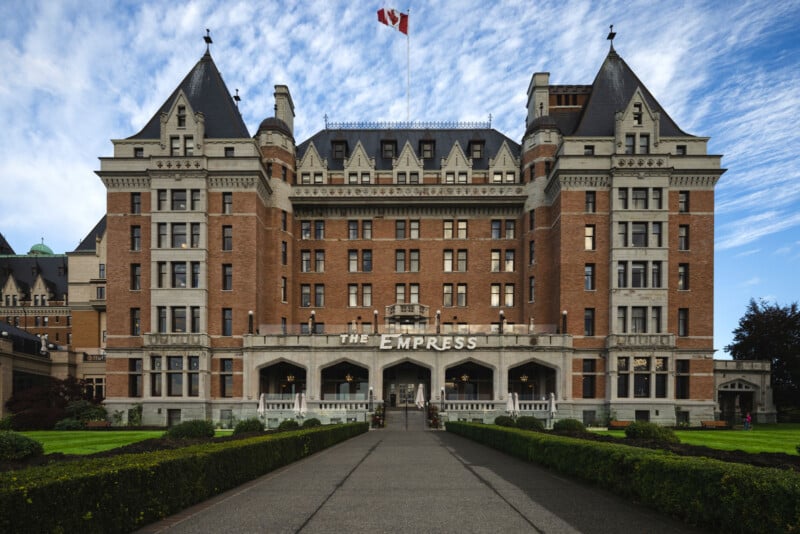 A grand historic hotel with turrets and a Canadian flag on top, labeled "The Empress," stands against a blue sky, framed by manicured gardens and a walkway leading to its main entrance.
