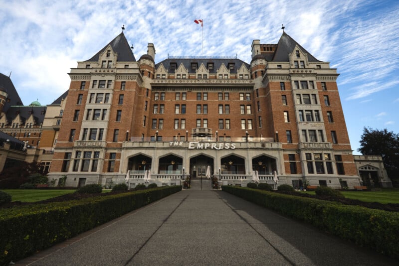 A grand, historic brick hotel with turrets and a Canadian flag on top, labeled "The Empress," stands at the end of a wide walkway, surrounded by manicured gardens under a blue sky with clouds.