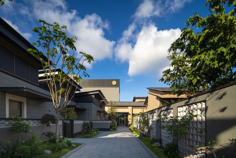 A modern Japanese-style building complex with tiled roofs, landscaped gardens, and a paved walkway, framed by green trees under a bright blue sky with scattered clouds.