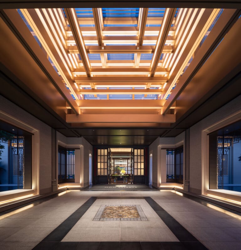 A modern, elegant hallway with a high, illuminated wooden ceiling, symmetrical lines, tiled floor, and large windows on both sides, leading to a distant, well-lit room with decorative art.