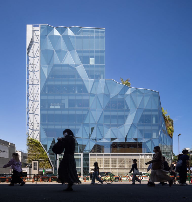 People walk in silhouette in front of a modern glass building with geometric, angular patterns on its facade, under a clear blue sky. The building has multiple levels, creating a dynamic, architectural design.