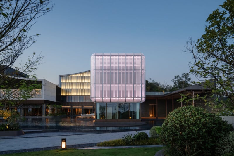 A modern building with a glowing, translucent cube structure at its center, surrounded by water and landscaped greenery, photographed at dusk with lights illuminating the architecture.