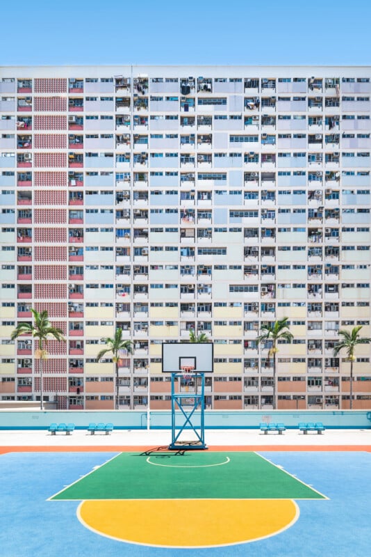 A vibrant outdoor basketball court stands in front of a tall, colorful apartment building with balconies, laundry, and four palm trees lining the court under a clear blue sky.