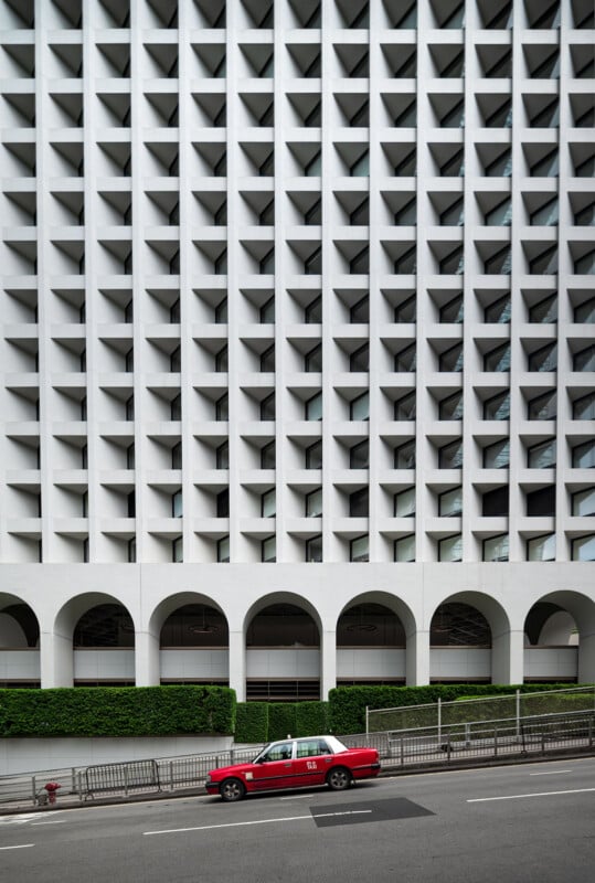 A red and white taxi drives past a modern building with a geometric facade of repeating square patterns and arched openings on the ground floor. Shrubs line the building’s base, and the street is mostly empty.