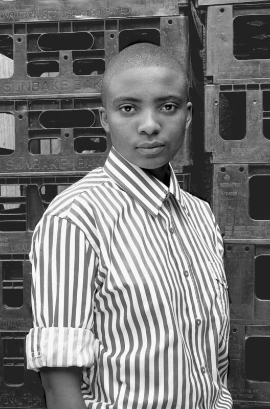 A person with short hair wearing a striped button-up shirt stands in front of a stack of plastic crates. They look directly at the camera with a neutral expression. The photo is in black and white.