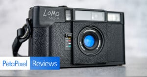 A close-up of a black Lomo LC-A analog camera with a lens and textured grip. The PetaPixel Reviews logo appears in the bottom left corner. The background is softly blurred.