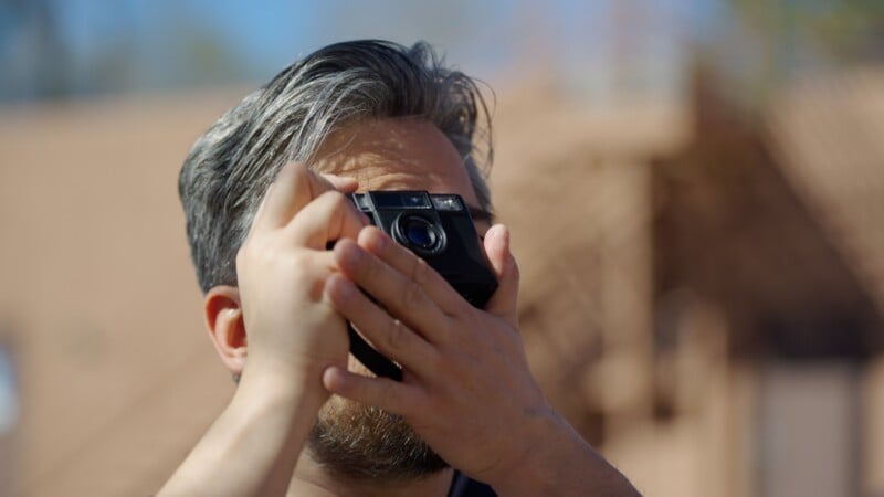 A person with gray-streaked hair holds a camera up to their face, preparing to take a photo outdoors on a sunny day. The background is blurred, emphasizing the person and the camera.