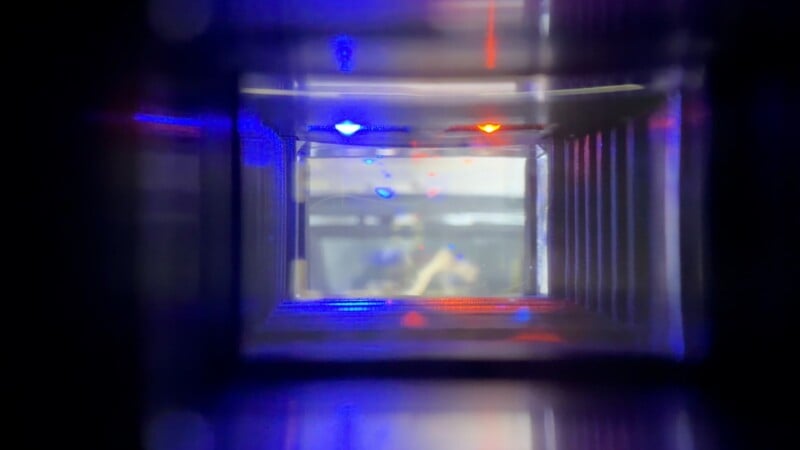 View through a rectangular tunnel with reflective surfaces, illuminated by blue and red lights. The tunnel leads to a blurred background, possibly showing people or objects in a laboratory or industrial setting.