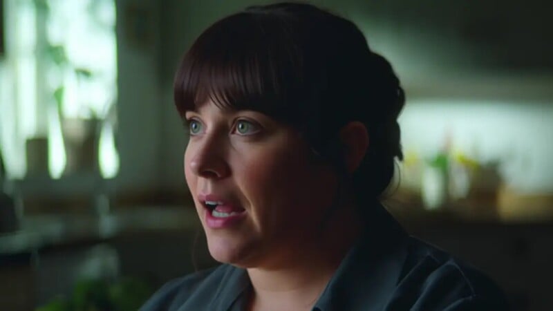 A woman with dark hair and bangs is speaking indoors. She has light eyes and is wearing a dark shirt. The background is softly lit, with some plants and kitchen items visible.