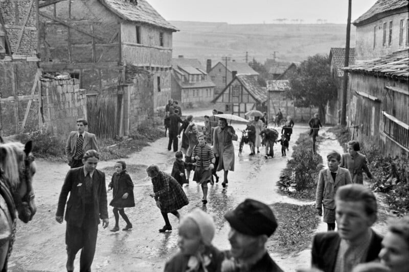 Black-and-white photo of people, including children and adults, walking up and down a wet, muddy street in a village with old stone buildings. Some carry umbrellas and wear coats, suggesting rainy weather.