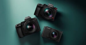 Three black Panasonic Lumix digital cameras are arranged on a green surface, with two mirrorless models and one compact model, all facing upward, displaying their prominent lenses.