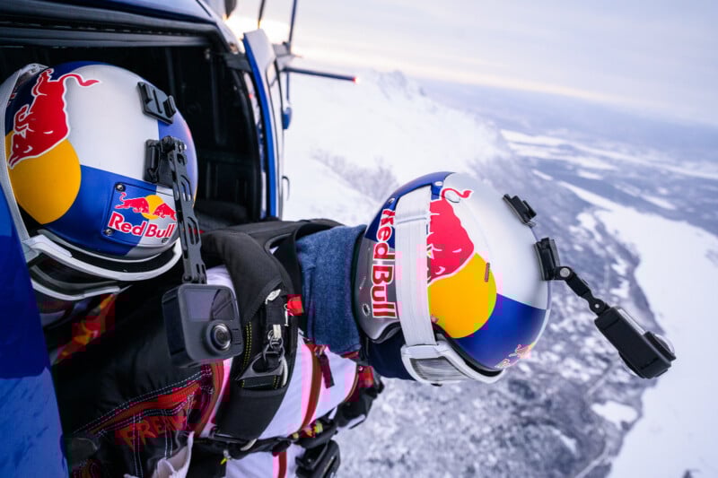 Dos paracaidistas con cascos y monos de Red Bull se preparan para saltar desde un helicóptero y volar sobre una zona montañosa nevada, ambos con cámaras montadas en sus cascos.