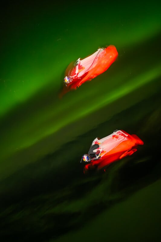 Por la noche, dos personas reman en un kayak rojo brillante sobre el agua verde oscuro, iluminada por luces vívidas, creando reflejos brillantes y un efecto abstracto surrealista.