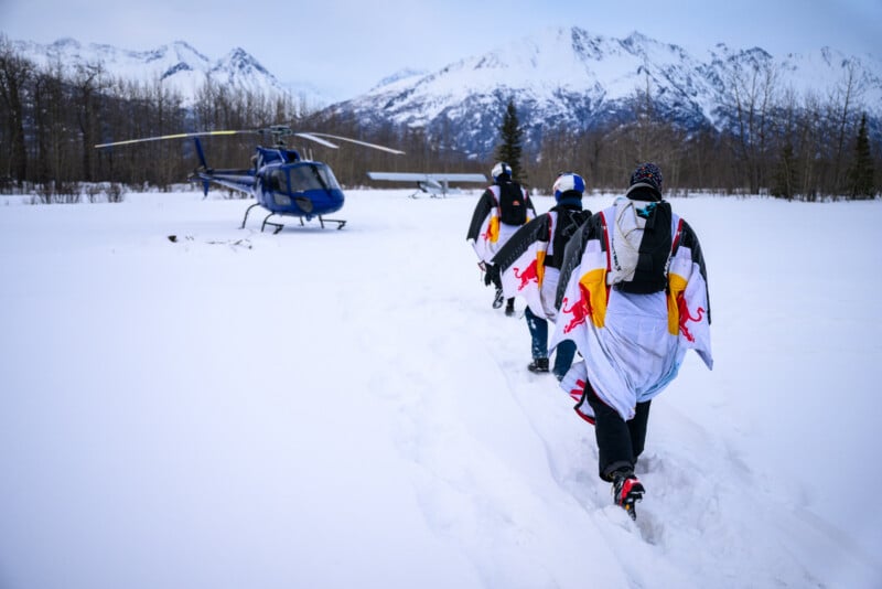 Tres hombres con trajes aéreos caminan por la nieve hacia un helicóptero azul, con montañas nevadas y árboles desnudos al fondo.