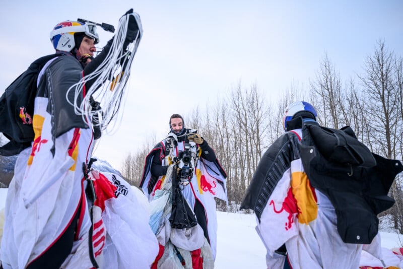 Tres personas con trajes aéreos adornados con el logo de Red Bull se encuentran en la nieve, sosteniendo paracaídas y equipo, contra un fondo de árboles desnudos y un cielo pálido.