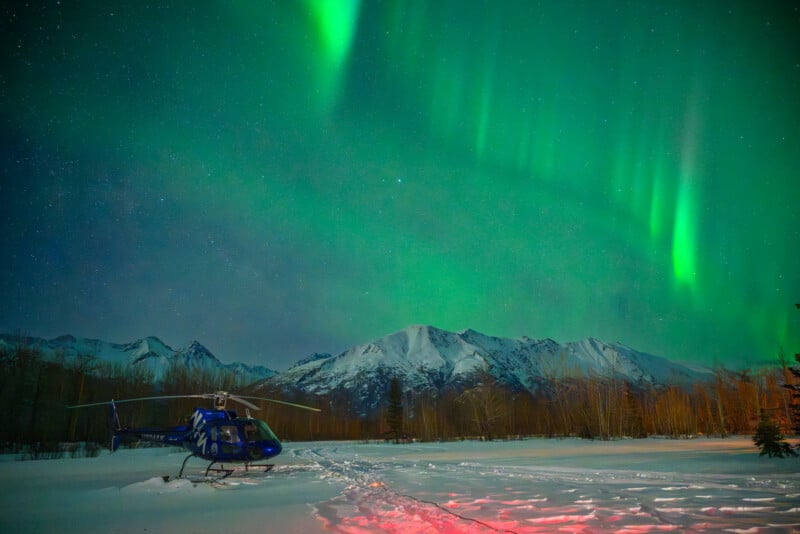 Un helicóptero azul está estacionado en la nieve, con la aurora boreal verde brillando en el cielo nocturno, con un telón de fondo de montañas cubiertas de nieve y árboles desnudos.