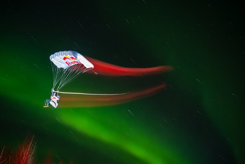 Un paracaidista que lleva un paracaídas con la marca Red Bull se desliza por el cielo nocturno, dejando un rastro de luz colorida sobre un fondo de estrellas y auroras boreales de un verde intenso.