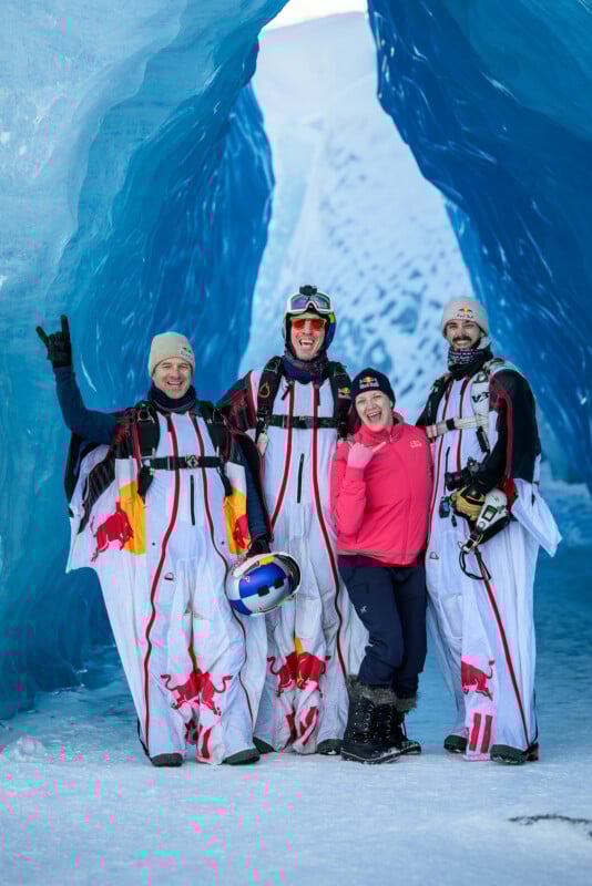 Cuatro personas vestidas con ropa para el frío posan en una cueva de hielo azul. Tres vestían trajes aéreos blancos con el logo de Red Bull y llevaban cascos, mientras que el cuarto, vestido con una camiseta rosa y pantalones negros, sonreía y hacía gestos con una mano.