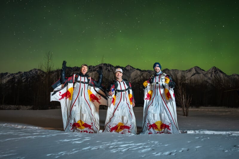 Tres personas vestidas con trajes aéreos con la marca Red Bull posan en la nieve por la noche, con las montañas al fondo y la aurora boreal verde iluminando el cielo sobre ellos.