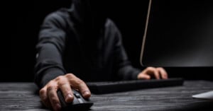 A person in a dark hoodie uses a computer mouse and keyboard at a desk, with their face obscured, suggesting anonymity or secrecy.