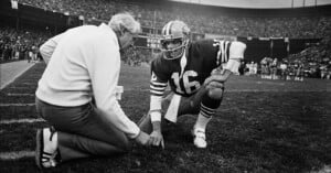 A football coach kneels on the sideline with a player in a number 16 jersey, both looking at a clipboard on the ground during a game in a packed stadium.