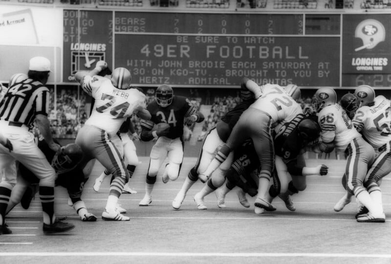 Black-and-white photo of a football game with a running back carrying the ball as players from both teams collide. A scoreboard and a "49er Football" promotion are visible in the background. Officials observe the play.