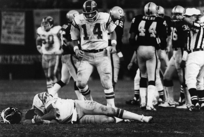 A football player lies on the ground clutching a football, while a teammate stands over him and opposing players stand nearby. The scene is tense and appears to be from an intense game.