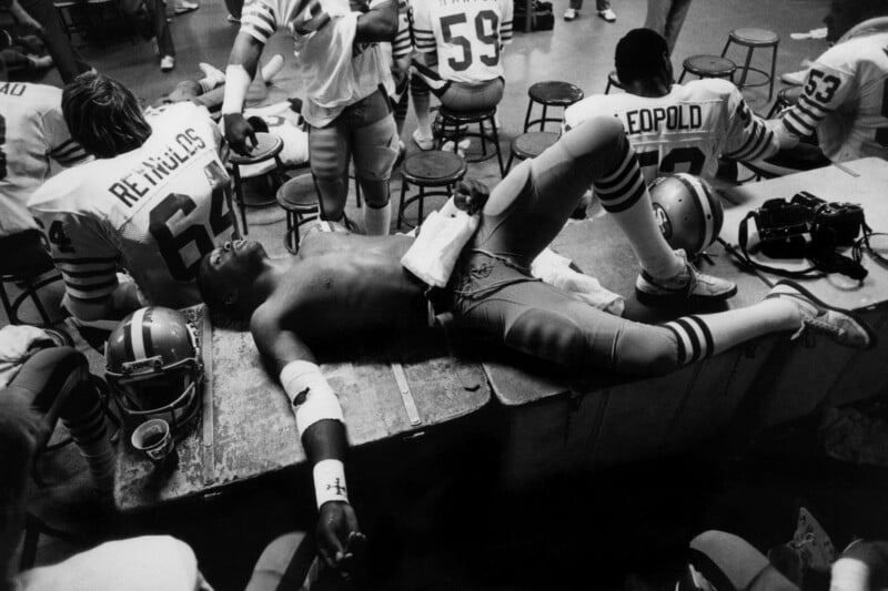 A shirtless football player lies on a table in a locker room, appearing exhausted, while teammates in uniforms stand or sit nearby. Helmets, jerseys, and equipment are scattered around.