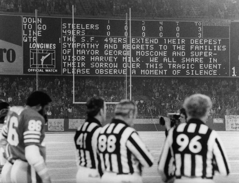 Black and white photo of a football field with players and referees standing still. The scoreboard displays a message offering condolences for the deaths of George Moscone and Harvey Milk, asking for a moment of silence.