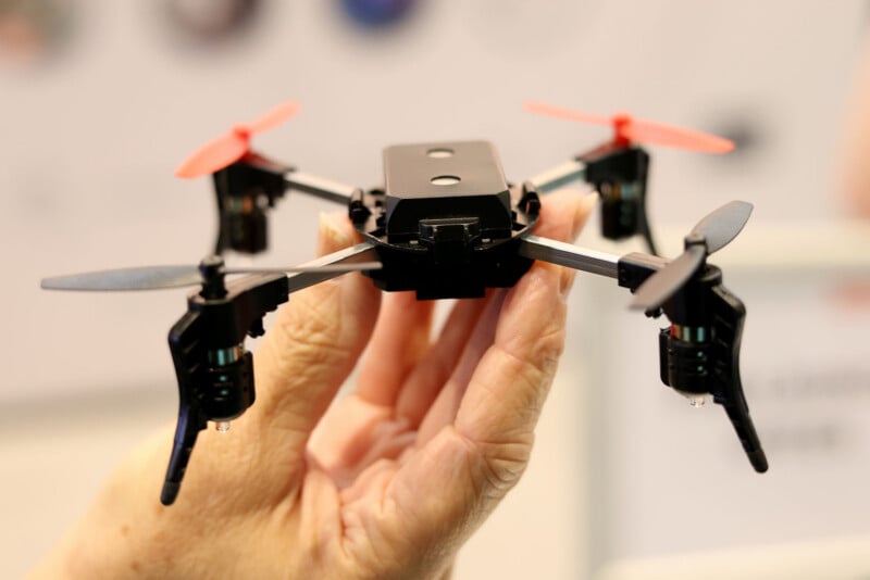 Una mano sostiene un pequeño dron cuadricóptero negro con dos hélices rojas y dos negras sobre un fondo borroso.