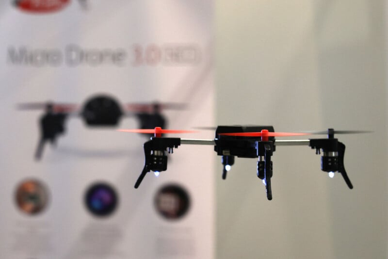 Un pequeño dron negro con hélices rojas vuela en el interior. El fondo está borroso, revelando un cartel blanco que dice "Micro UAV 3.0" Así como algunos íconos e imágenes.