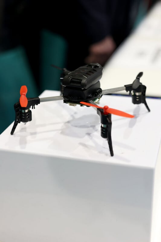 Un pequeño dron cuadricóptero negro con dos hélices naranjas y dos hélices negras se muestra sobre una base blanca con un fondo borroso.