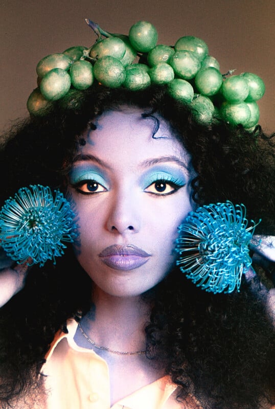 A person with curly hair wears a crown made of green grapes and holds two bright blue, spiky decorative spheres near their face. They have bold teal eyeshadow and a neutral expression.