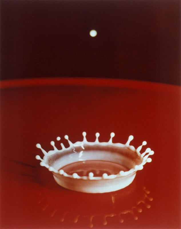 A close-up photo of a white milk drop creating a crown-shaped splash pattern on a smooth red surface, with a single white dot floating above in the dark background.
