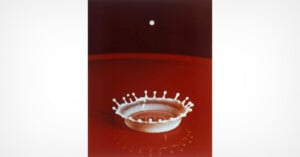 A close-up photo of a white milk drop splashing into a crown shape on a red liquid surface, with a single white droplet suspended above in midair against a dark background.