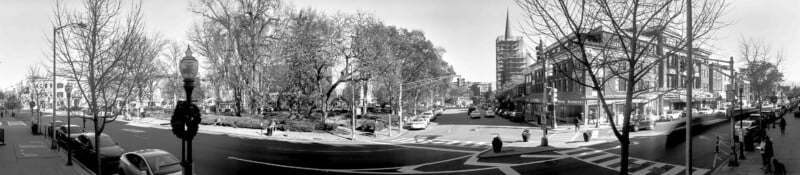 Amplio panorama en blanco y negro de una intersección de la ciudad con árboles, peatones, automóviles y varios edificios de varios pisos a lo largo de la calle. El cielo está despejado y las ramas desnudas enmarcan la escena.