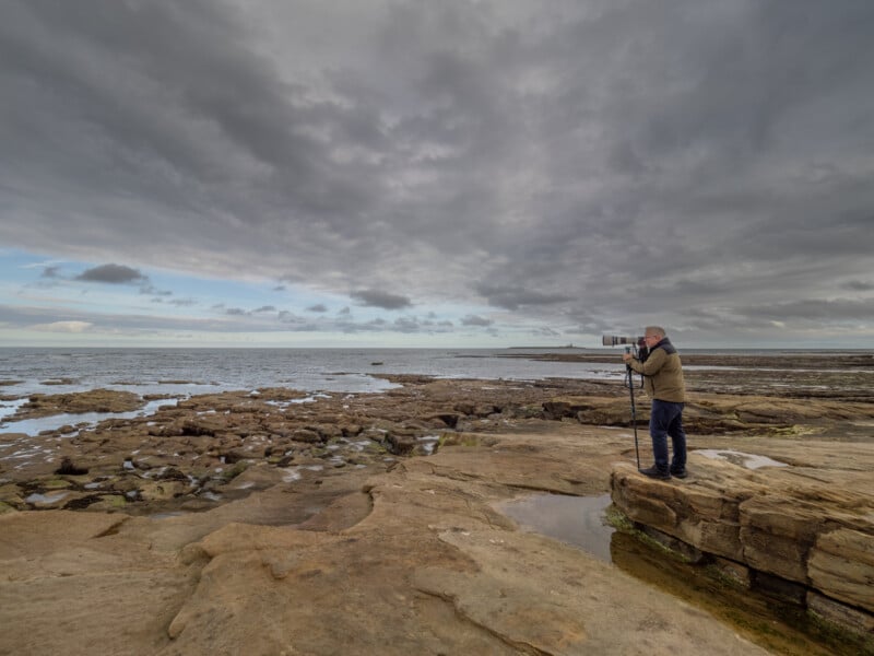 A person stands on rocky coastal terrain with a camera on a tripod, aiming it toward the cloudy sky and distant sea. The scene is expansive with dramatic clouds and scattered pools of water on the rocks.