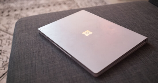 A closed silver Microsoft Surface laptop rests on a gray fabric couch, with a patterned carpet visible in the background.