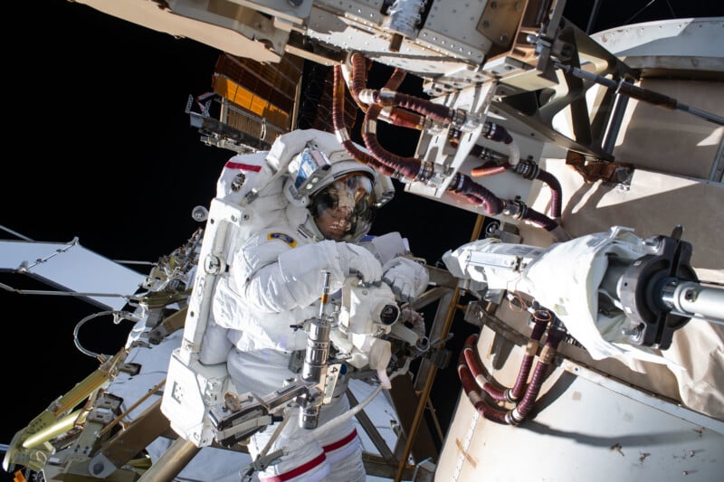 An astronaut in a spacesuit works outside the International Space Station, handling equipment and tools against a backdrop of space and station structures.