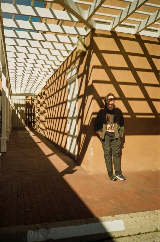 A person stands against a tan building, partially in shadow, under a white geometric pergola that casts bold square patterns of light and shadow on the wall and ground. The person wears sunglasses, a jacket, and dark pants.