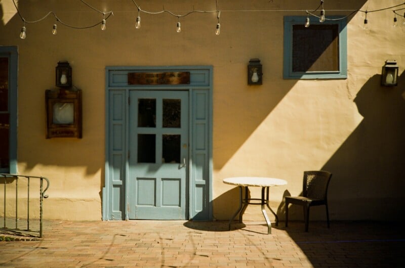 A sunlit patio with a light blue door, a small round table, and a single chair. String lights hang above, casting shadows on the beige wall and brick ground. Two windows and wall lamps are visible on the building.