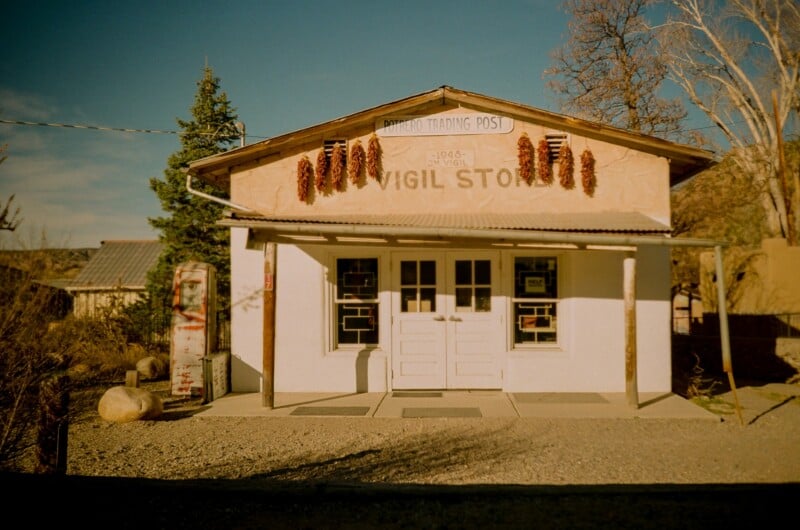 A small, rustic store with a white front, labeled "Vigil Store," decorated with red chile ristras hanging above the door, sits in bright sunlight with trees and shrubs around it.